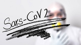 In the forefront, Sars-Cov2 written in black ink on a perspex screen. In the background, a person wearing forensic clothing.