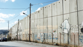 A tall concrete border wall surrounded by barbed wire and painted with graffiti.
