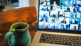 Work from home image. There is a laptop on a wooden table next to a blue mug. The laptop screen shows a group Zoom video call with the participants faces slightly blurred.