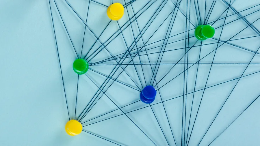 A network of black thread connects an array of colorful pushpins on a light blue background. The pins are red, yellow, green, and blue, creating a vibrant and eye-catching pattern. The threads create lines that crisscross and connect each pushpin to the others, forming a complex web. 