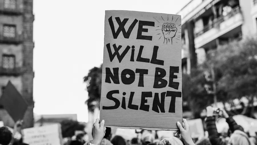 A black and white photograph depicts a crowd of people gathered in an urban setting, with many holding signs. The focal point is a sign in the foreground that reads, "WE WILL NOT BE SILENT," accompanied by a hand drawn fist symbol. A person's hands hold the sign aloft, while buildings and trees create the backdrop.