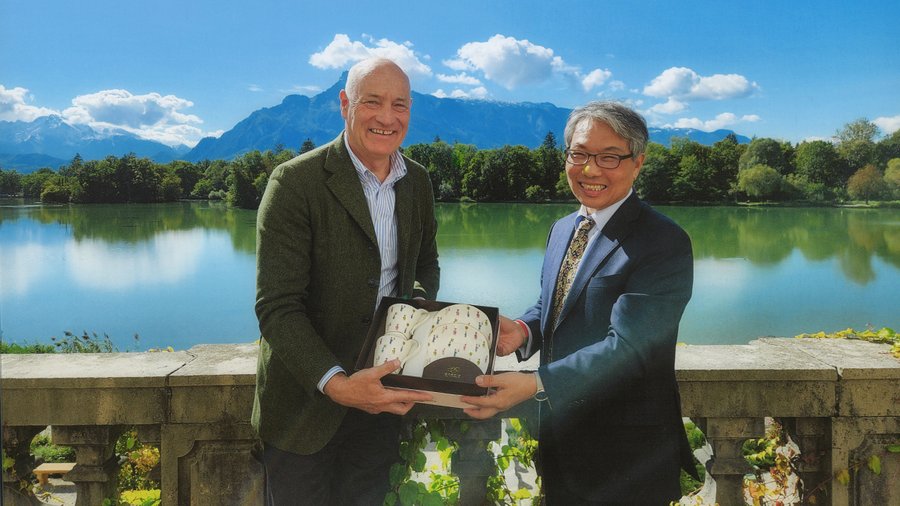 Two men stand on a balcony holding dishware with a mountain in the background