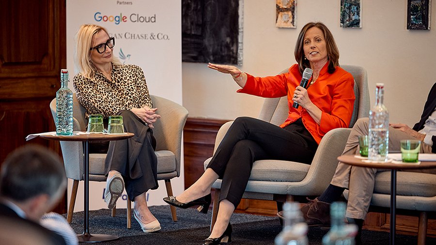 two women seated on a stage, with one woman in a red blouse speaking with her right arm extended.
