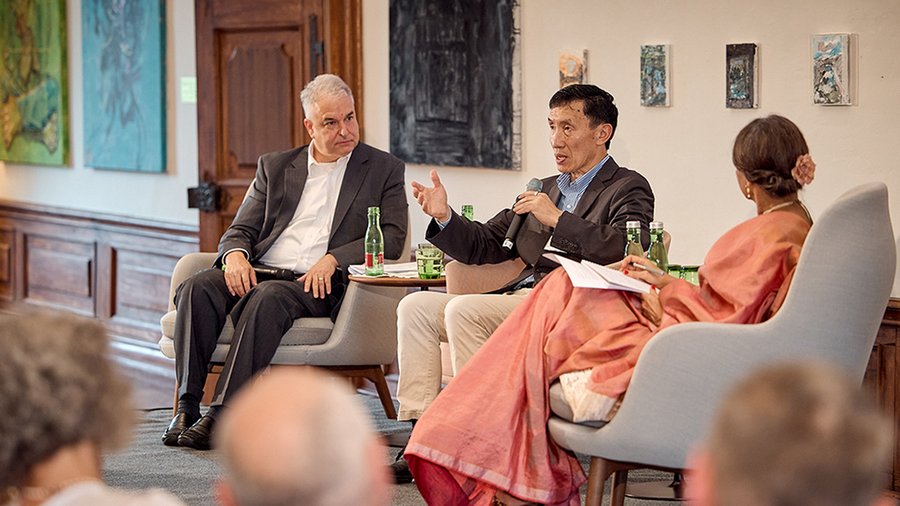 Three people are seated side by side on a stage. A man on the left looks attentively at the man in the center who is speaking into the microphone. A woman wearing a long dress is seated to the right of them with her back facing the camera. 