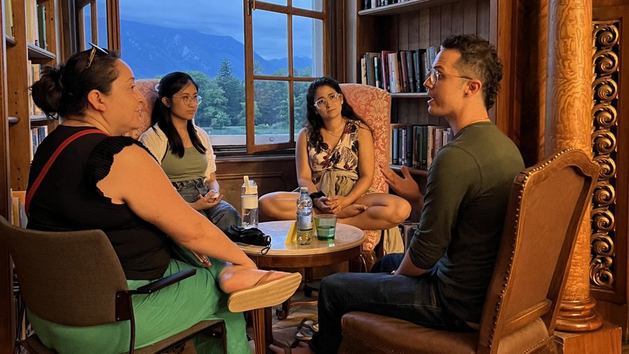 a group of people sit in a round circle surrounded by books and an open window with a mountain view