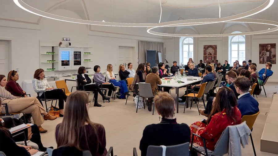 Inside a large room, many people sit in a circle of chairs with a smaller circle around a table in the middle.