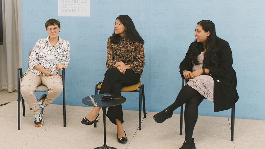 Three women sit on chairs and the one on the left is speaking