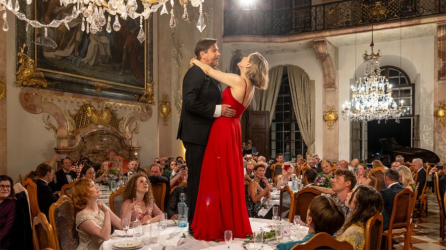 A man and a suit and a woman in a red dress stand on a table