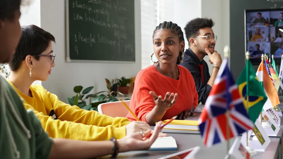 Four young adults are seated around a table in a classroom during daytime. One woman with braided hair and an orange shirt speaks while gesturing with her hands. Other students in the foreground wear a yellow shirt, and a green shirt. A man with glasses sits to the right, listening. The table has miniature flags on it, and there are tablets and notebooks. A chalkboard and plant are visible in the background. 