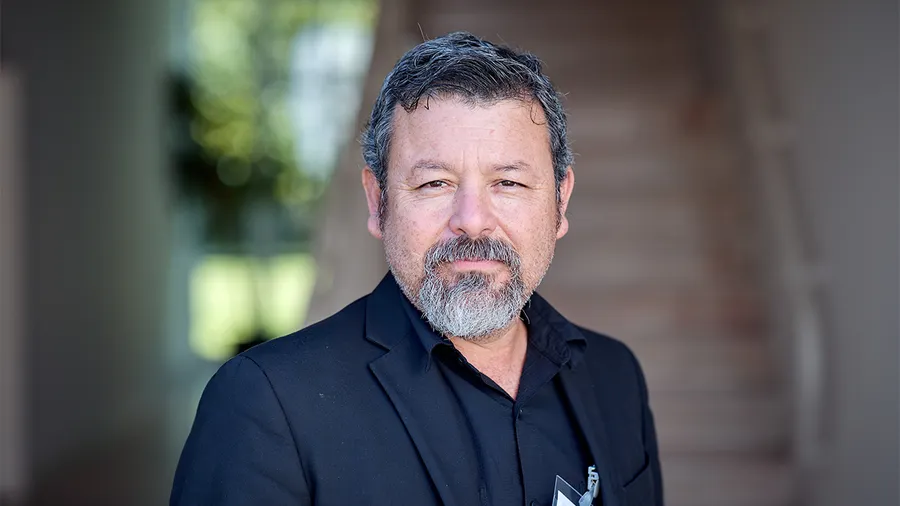 A man with dark hair stands looking directly into the camera