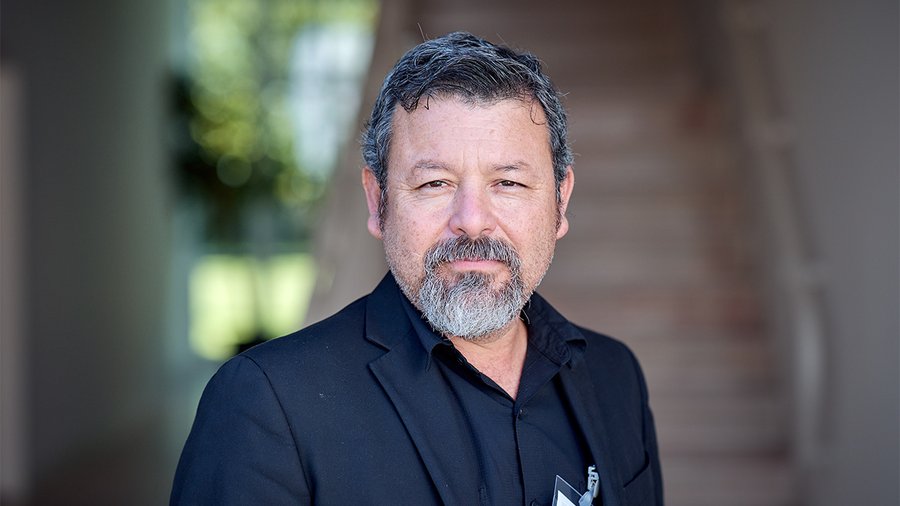 A man with dark hair stands looking directly into the camera