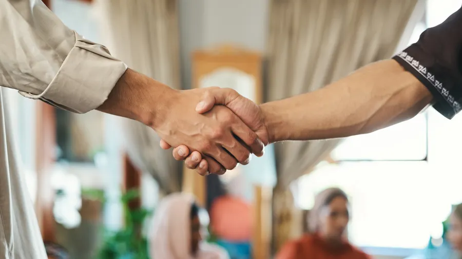 A close up shot shows two adults shaking hands. In the background, a group of people wearing head coverings and casual clothing gather indoors. The photo is brightly lit with natural light and neutral interior design
