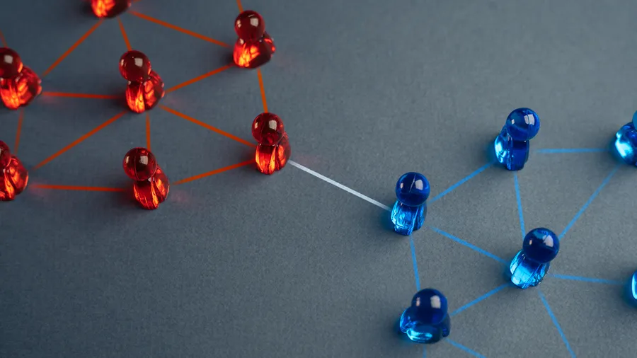A network communication concept is represented by shiny push pins that are connected with string. Red and blue pins are on opposite sides of the image, with connecting red and blue lines. The pins are set against a textured gray surface. 