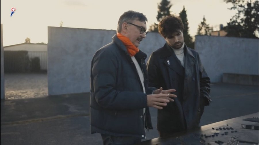 Two men stand outside on a sunny day, facing a table that has figurines. 