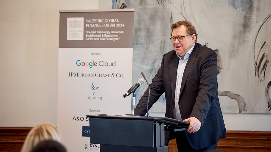 a man with glasses wearing a suit stands at a podium speaking into a microphone