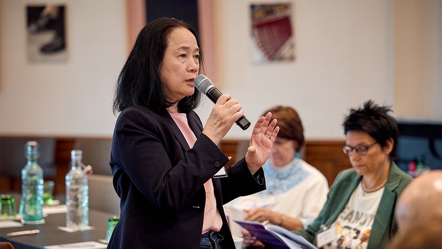 A woman in a pink shirt and black blazer speaks into a microphone while turning to the right.