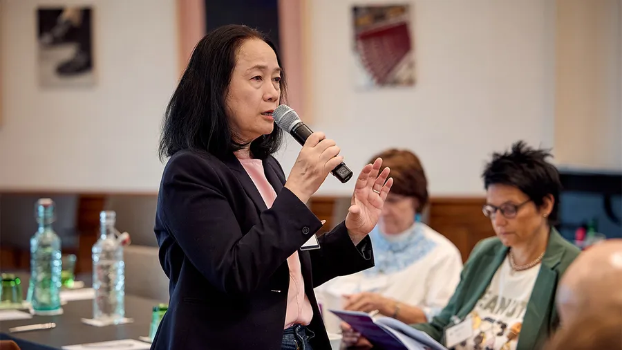 A woman in a pink shirt and black blazer speaks into a microphone while turning to the right.
