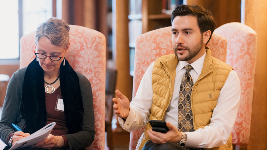 Amr, a man with dark hair wearing a yellow vest, speaks next to a woman who looks down while taking notes