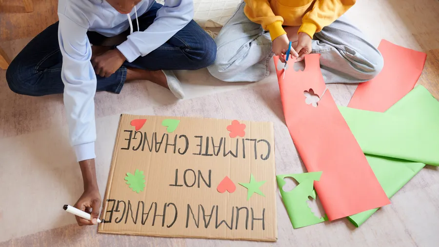 Two people are visible in a bright, eye-level shot inside of a home. Both are sitting on the floor, and one is cutting red paper with scissors. Next to them is a piece of cardboard with text that says, "Human Change NOT Climate Change." One person is holding a black marker. Some scraps of red and green paper are next to the sign.