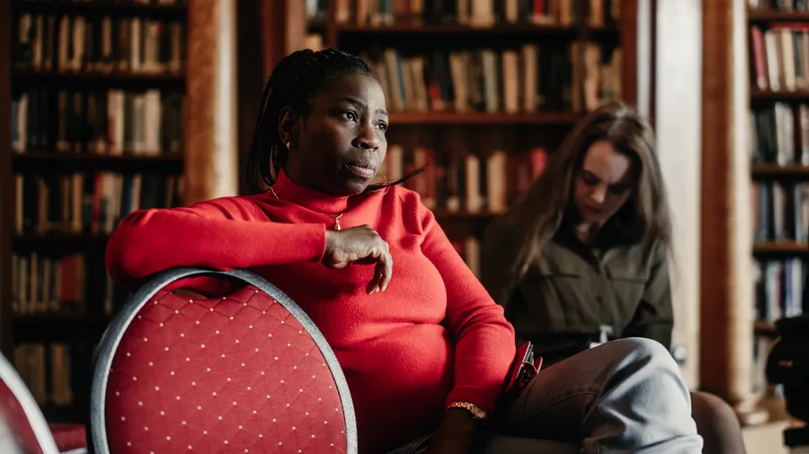 a woman wearing a striking red sweater sits in the library with her arm resting on a chair