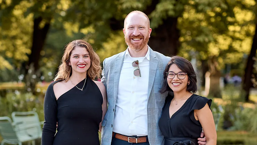 Three people stand smiling next to each other in front of an outdoor background.