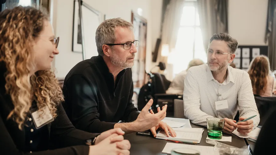 three people sit at a table having a conversation, with the man in the middle speaking