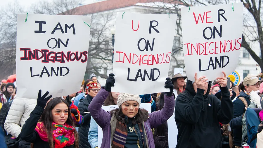 three indigenous people marching with a crowd, and each holding a sign