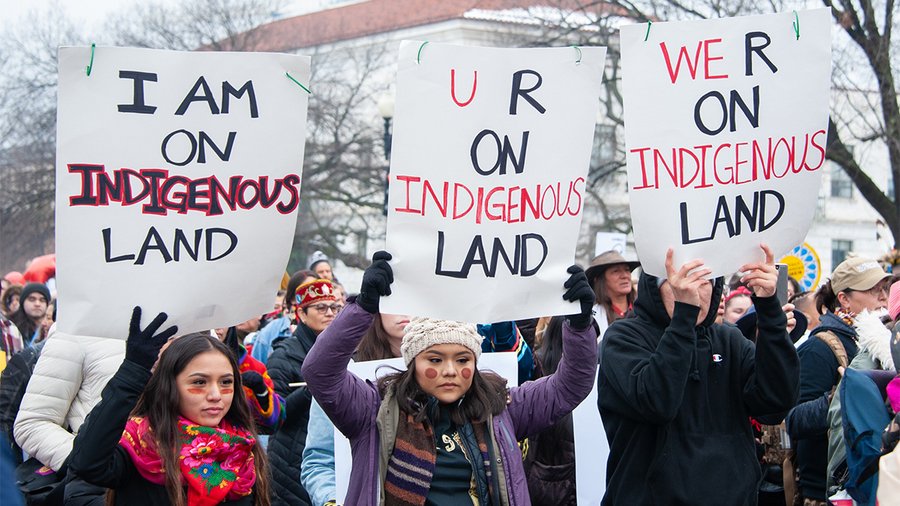 three indigenous people marching with a crowd, and each holding a sign