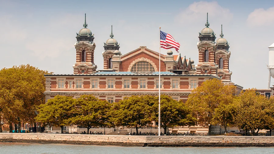 A photo of the main immigration on Ellis Island in New York harbor with trees and an American flag in the foreground.