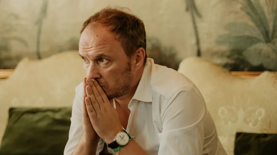 Petrit Selimi, a man in a white, sits with a contemplative look on his face and his hands together near his mouth.