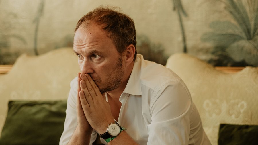 Petrit Selimi, a man in a white, sits with a contemplative look on his face and his hands together near his mouth.