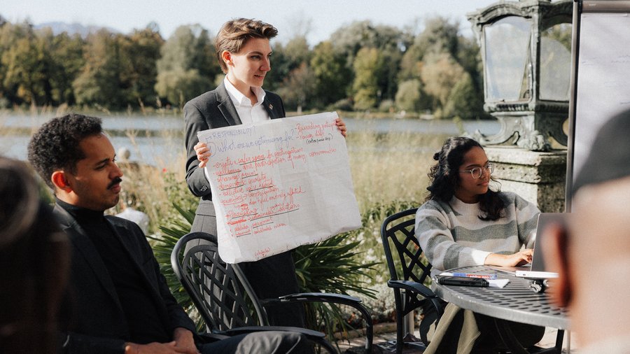 Three individuals sit outside, with one standing in the middle holding a poster with writing on it.