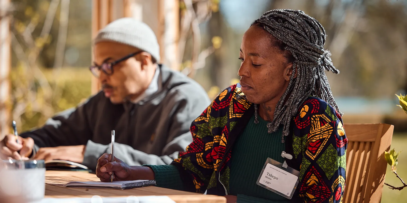 a woman sits outside at a table writing on a paper