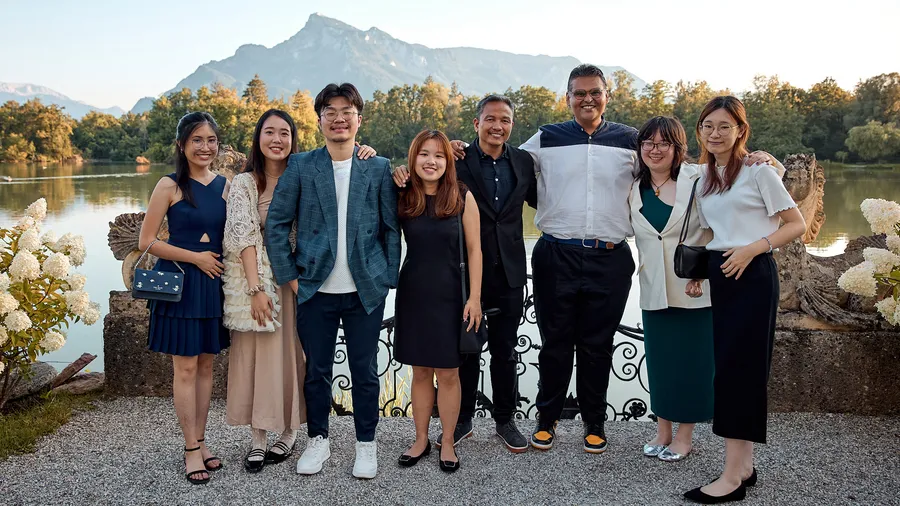 a group of 8 individuals stand posing and smiling for the camera with a mountain and lake in the background.