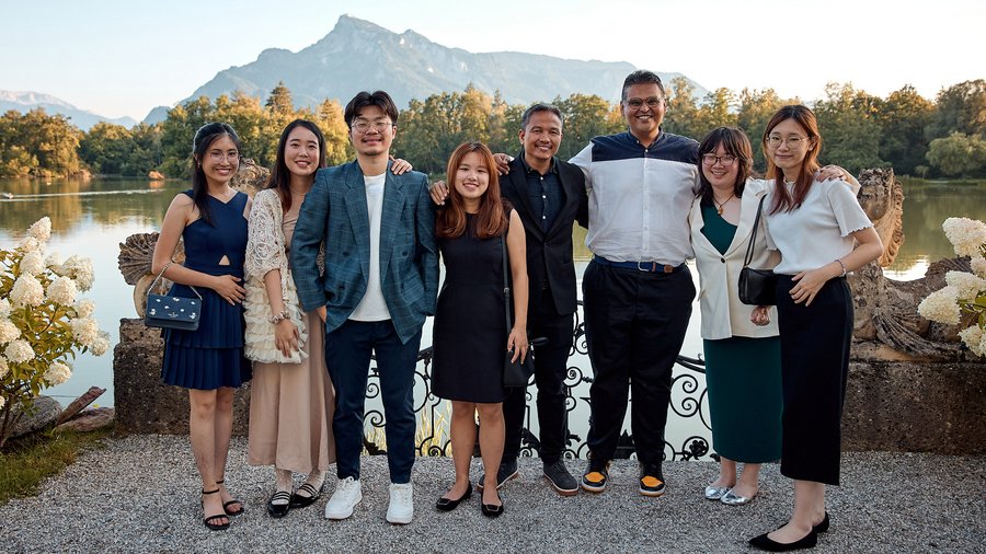 a group of 8 individuals stand posing and smiling for the camera with a mountain and lake in the background.