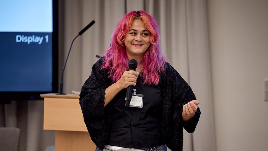 a woman wearing a dark shirt with colored hair holds a microphone and speaks