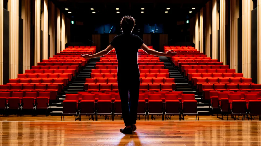 An actor stands on a stage facing an empty auditorium, with his back turned to the camera.