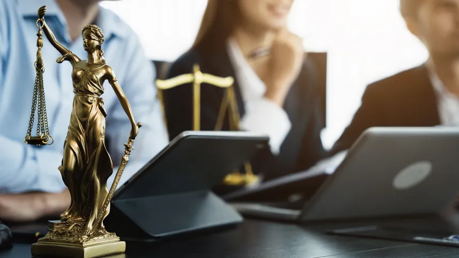 A shiny gold-colored statue of Lady Justice stands prominently on a desk, complete with scales and sword. Behind it, three partially visible people, likely adults, are seated at a table, two with laptops open. The setting appears to be a modern office with a professional atmosphere. The lighting is bright, enhancing the polished surfaces. 