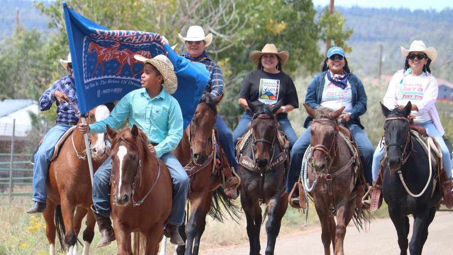 A group of Native American riders on horseback participate in a voter mobilization event, with a young rider at the front holding a blue flag that reads, "Your Vote is the Reins of Democracy," as they move along a rural road