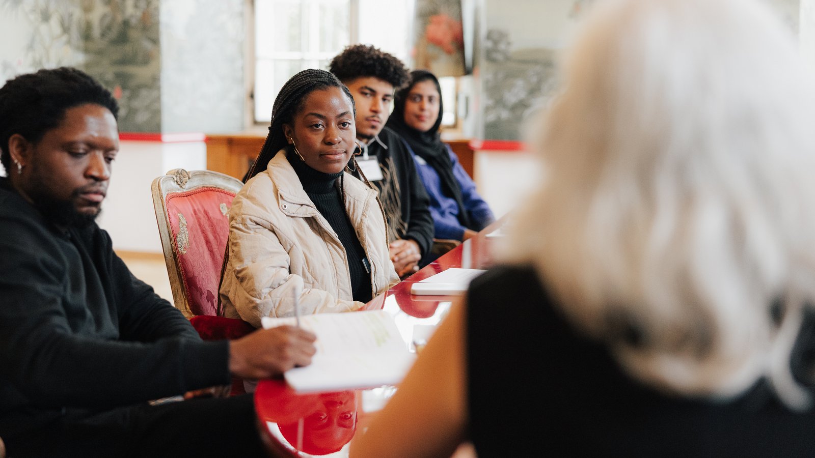 A group of young people sit at a table listening attentively to a woman in the forefront.