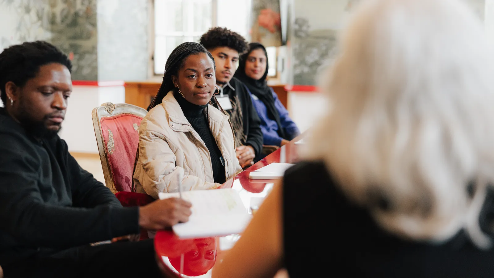 A group of young people sit at a table listening attentively to a woman in the forefront.