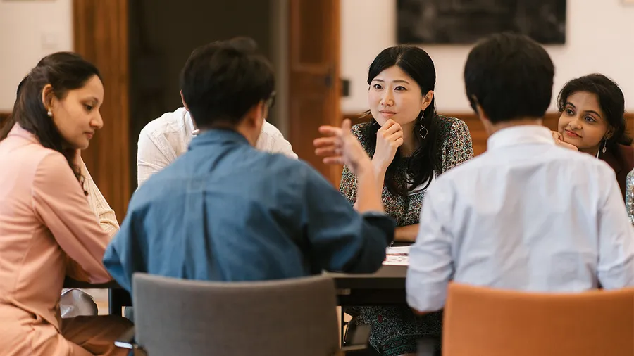 a group of six people sit around a table, with the backs of two men to the camera and the focus on a young Japanese woman
