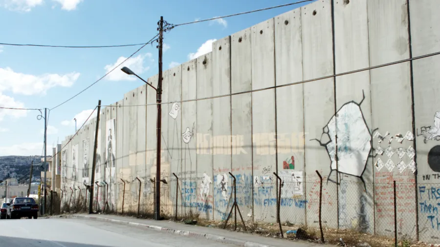 A tall concrete border wall surrounded by barbed wire and painted with graffiti. 