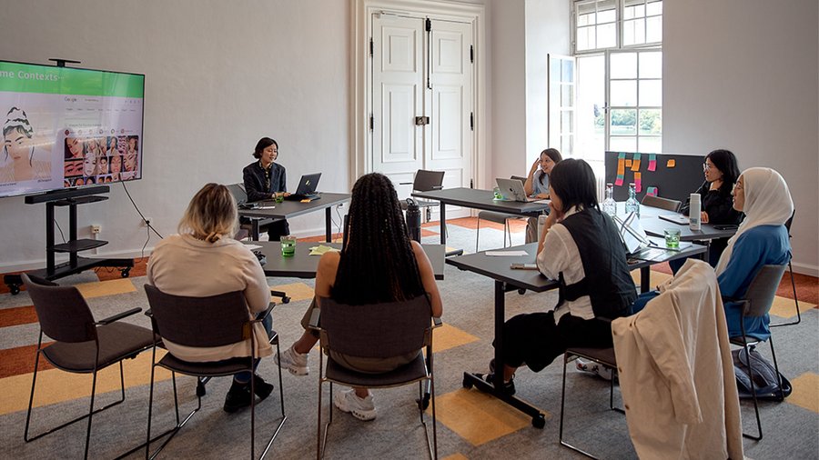 a circle of young women seated at desks listen to a presentation which is shown on a TV