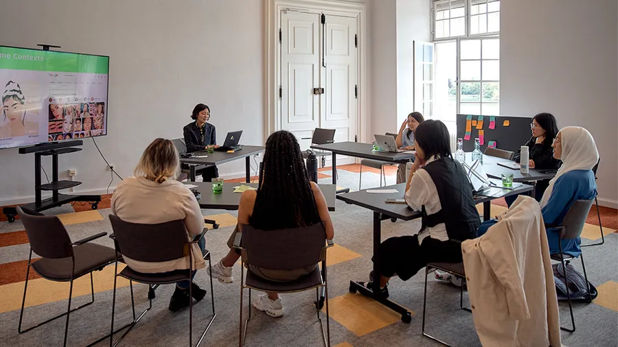 a circle of young women seated at desks listen to a presentation which is shown on a TV