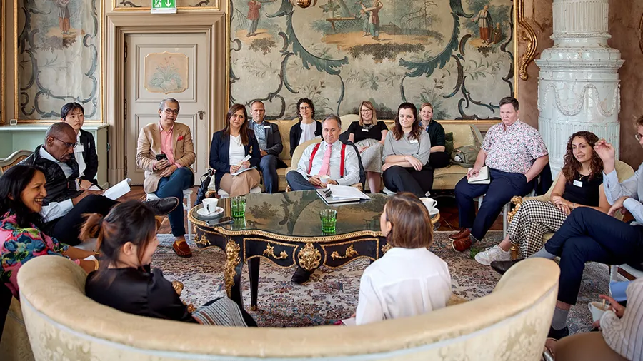 A group of people are seated in a circle in the Green Salon and attentively watch two female speakers.