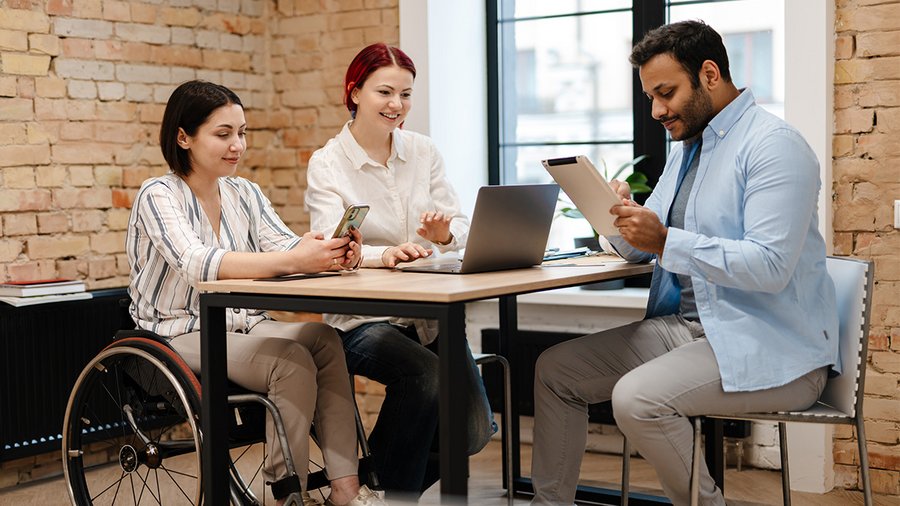 Three people sit around a desk, as one woman in a wheelchair looks at a phone, another woman uses a computer, and a man writes notes