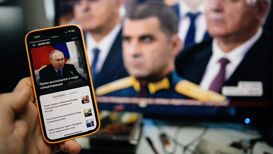A hand holds a phone with Russian news with a TV screen in the background