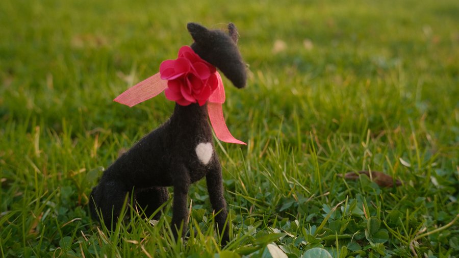 Needle felted symbol of Lucy Like-a-Charm, a black greyhound dog, wearing a pink ribbon, sitting in the grass, at the Schloss Leopoldskron.