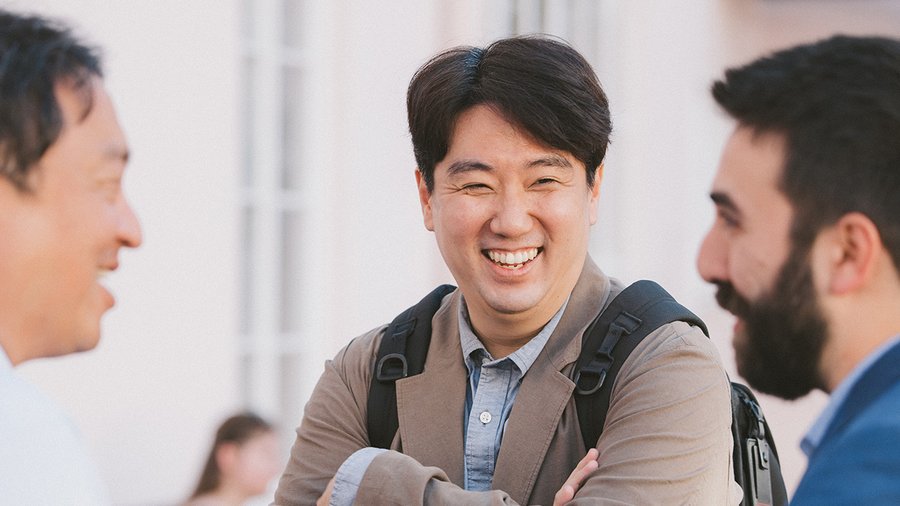 A man with dark hair laughs and looks towards a man to the right who is not fully visible